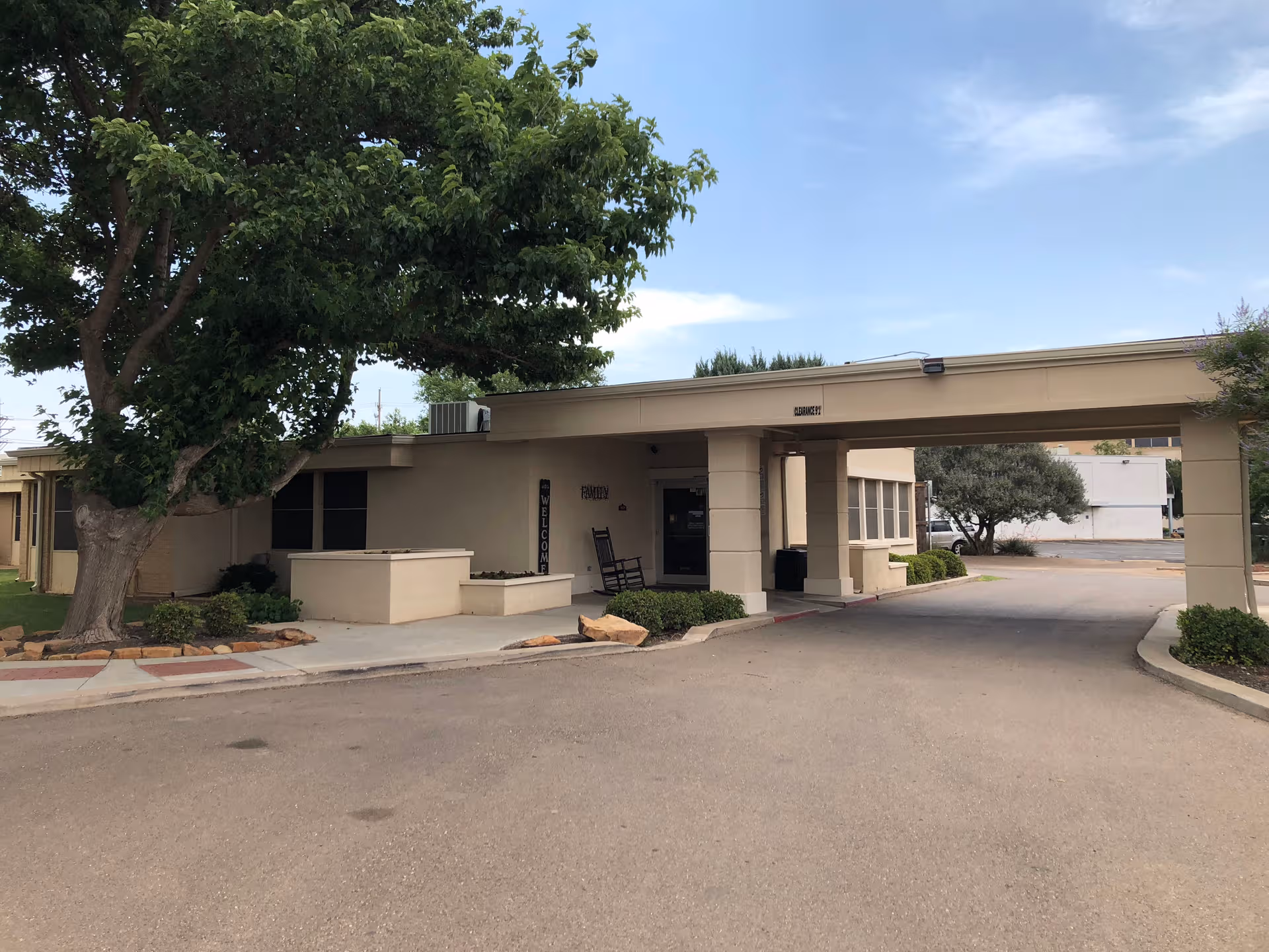 Exterior view of a single-story senior care facility entrance with driveway and a tree.