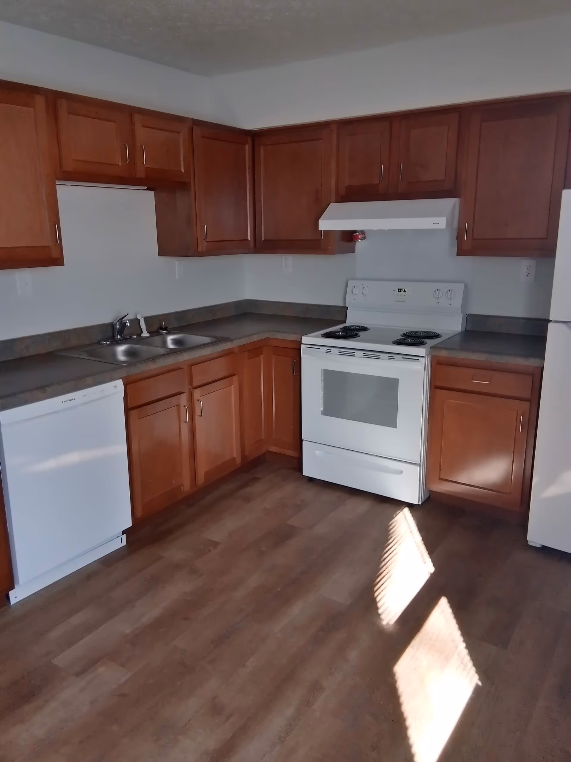 L-shaped kitchen with brown cabinets, white stove, sink, and dishwasher.