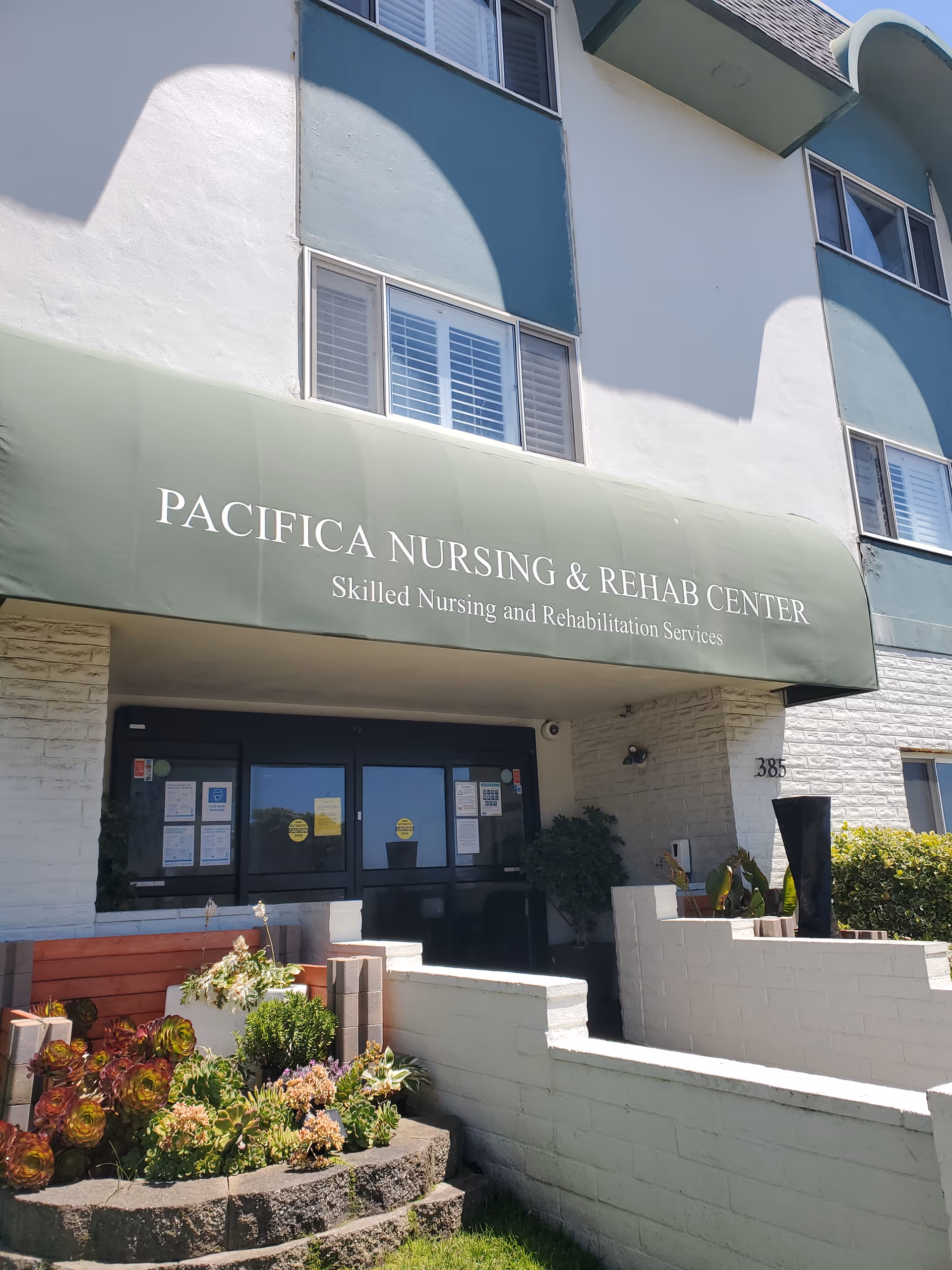 Exterior view of Pacifica Nursing and Rehab Center entrance with green awning and plants.