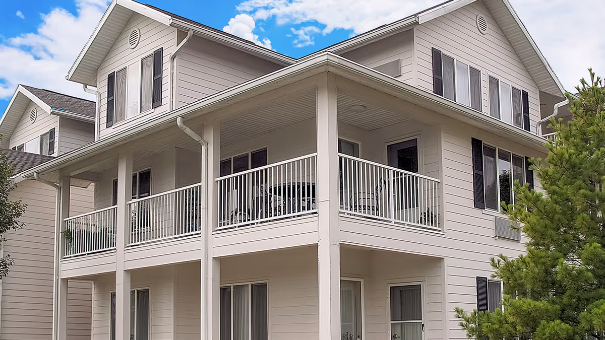Exterior view of a two-story residential building with beige siding, multiple windows with black shutters, and a wrap-around balcony with white railings. The sky is blue with some clouds and there are green trees around the building.