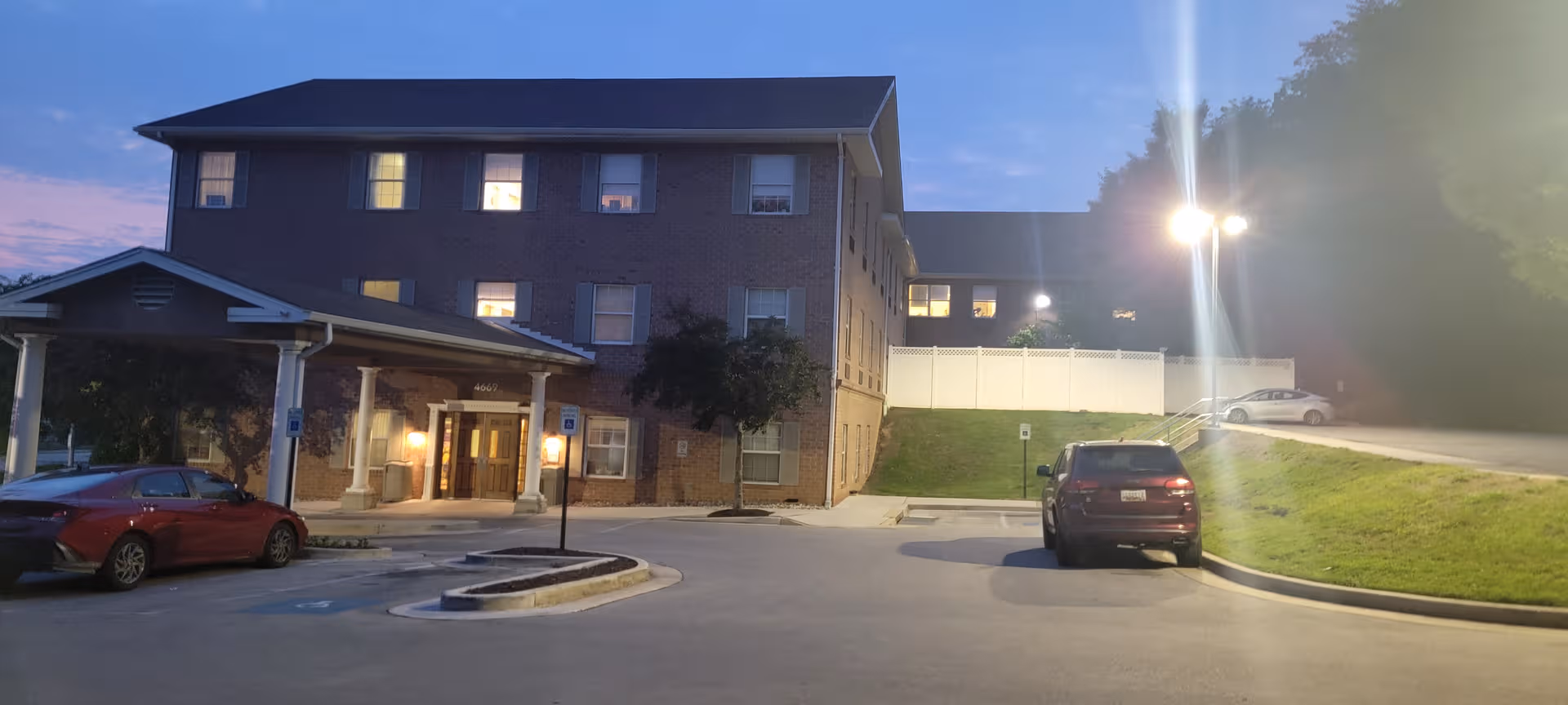 Brick four-story senior care building with a driveway, parking lot, and streetlights at dusk.