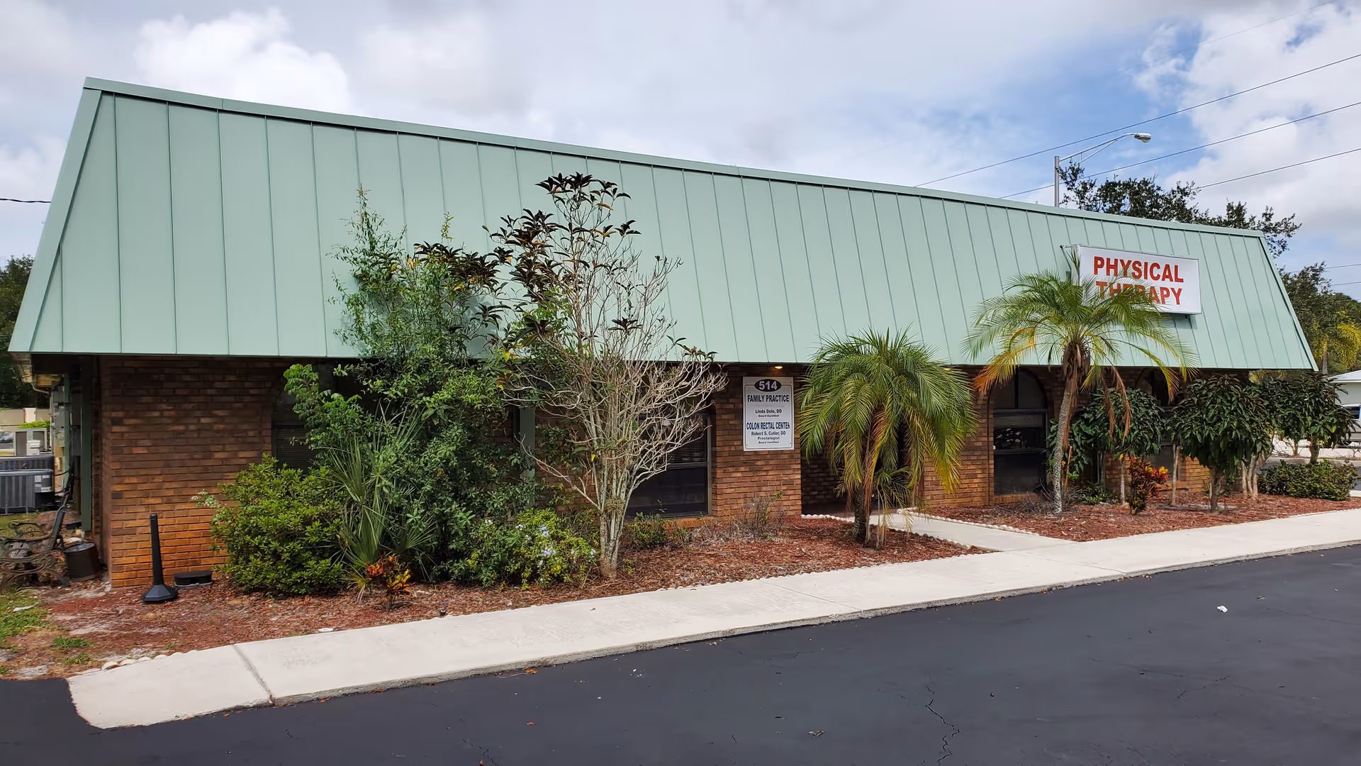 Brick storefront-style building with green metal roof, landscaped frontage, and a Physical Therapy sign.