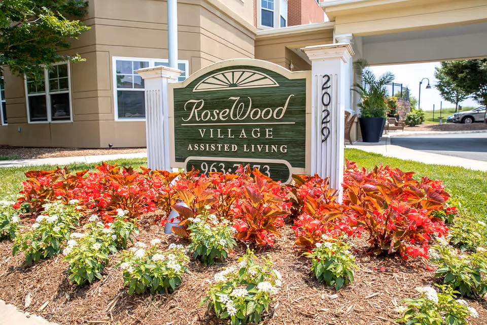 Wooden sign reading 'RoseWood Village Assisted Living' surrounded by red and white flowers at the facility entrance.