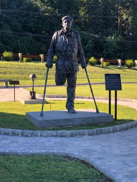 Bronze statue of a man with crutches on a pedestal in a park-like outdoor area.