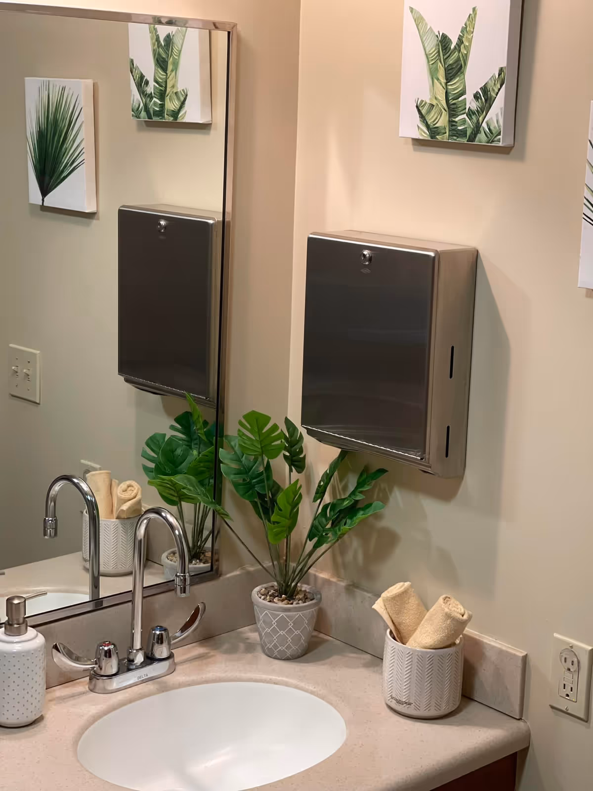 Bathroom counter with sink, mirror, metal dispenser, and decorative plants at Wildwood Care Center.