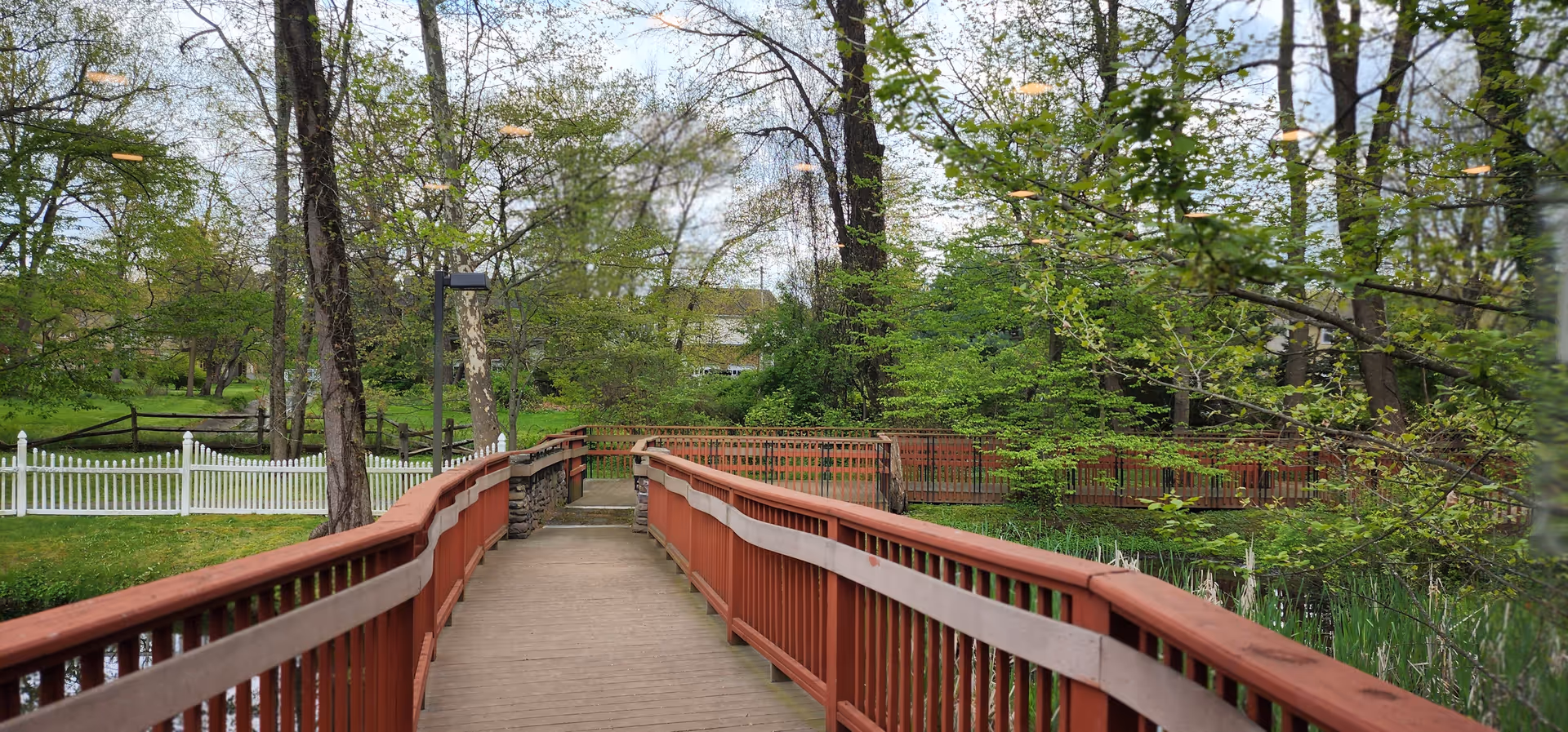 A red wooden bridge spans a stream in a lush, tree-filled outdoor area.