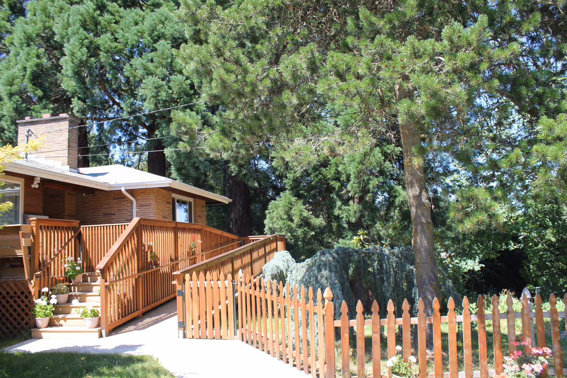 Small brick building with wooden ramp, stairs, and fenced yard amid tall trees.