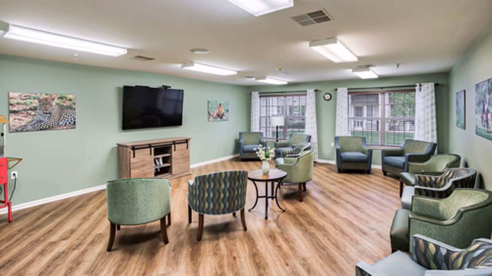 Green-walled common lounge with chairs, tables, and a wall TV.