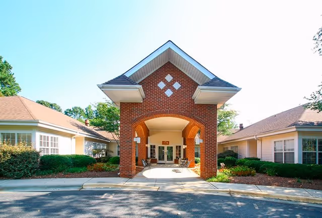 Front exterior view of Carriage House Senior Living Community featuring a brick entrance with an arched covered walkway, flanked by single-story buildings with beige siding and surrounded by greenery.