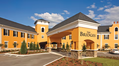 Exterior view of a large yellow building with a covered entrance labeled 'The Bridges'. The building has multiple windows, a tower-like structure, and a parking area with a few cars. The sky is blue with some clouds.