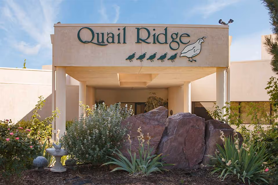 Entrance of Quail Ridge Assisted Living & Memory Care facility featuring a beige building with a covered entryway. The facility's name 'Quail Ridge' is displayed prominently above the entrance along with a graphic of a quail and five smaller birds. In front of the entrance, there are large rocks and various plants and shrubs.