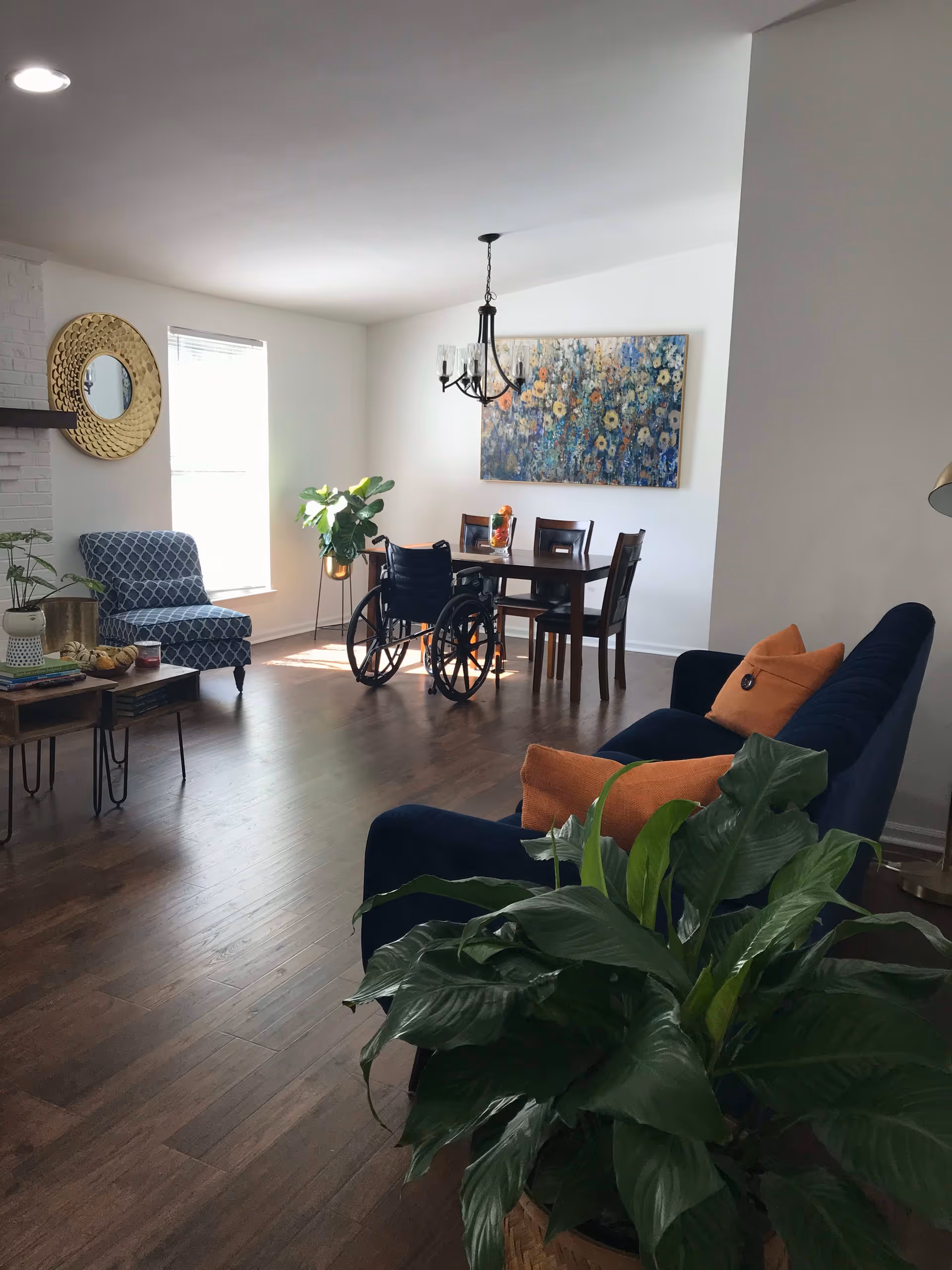 A bright living and dining area with wooden flooring. The living area has a navy blue sofa with orange cushions and a large green plant in the foreground. There is a blue patterned armchair near a window with blinds and a round gold-framed mirror on the wall. The dining area features a wooden table with four chairs, a wheelchair positioned at the table, a chandelier overhead, and a colorful floral painting on the wall.