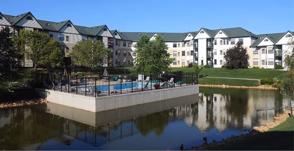 Exterior view of Autumn Woods facility with a fenced pool overlooking a pond and lawns.