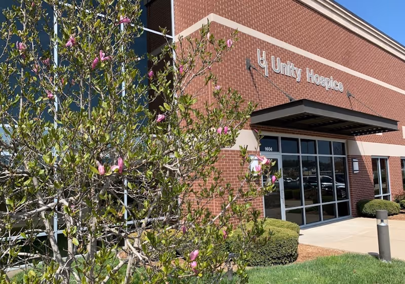 Brick Unity Hospice building entrance with glass doors and flowering shrubs in foreground.
