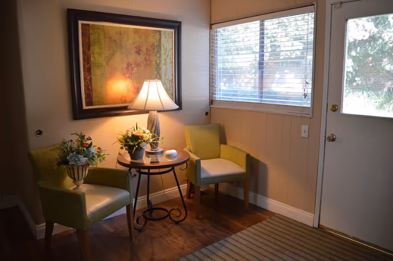 A cozy corner in a senior living facility with two green armchairs around a small round wooden table. The table holds a lamp with a white shade and two flower arrangements. A large framed artwork hangs on the wall behind the lamp. A window with blinds and a door with a window panel are visible on the right side. The floor is wooden with a striped rug near the door.