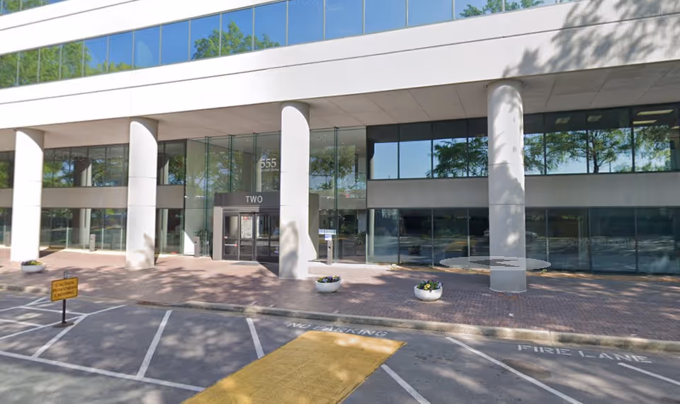 Modern glass-front building entrance with pillars, parking lot, and decorative planters.