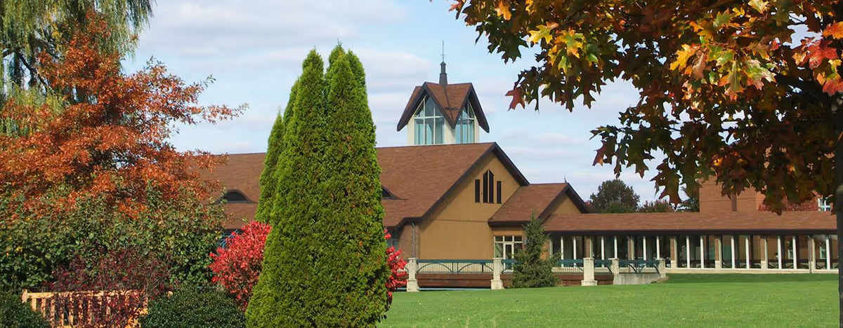 A large building with a brown roof and beige walls surrounded by green grass and various trees with autumn foliage under a partly cloudy sky.