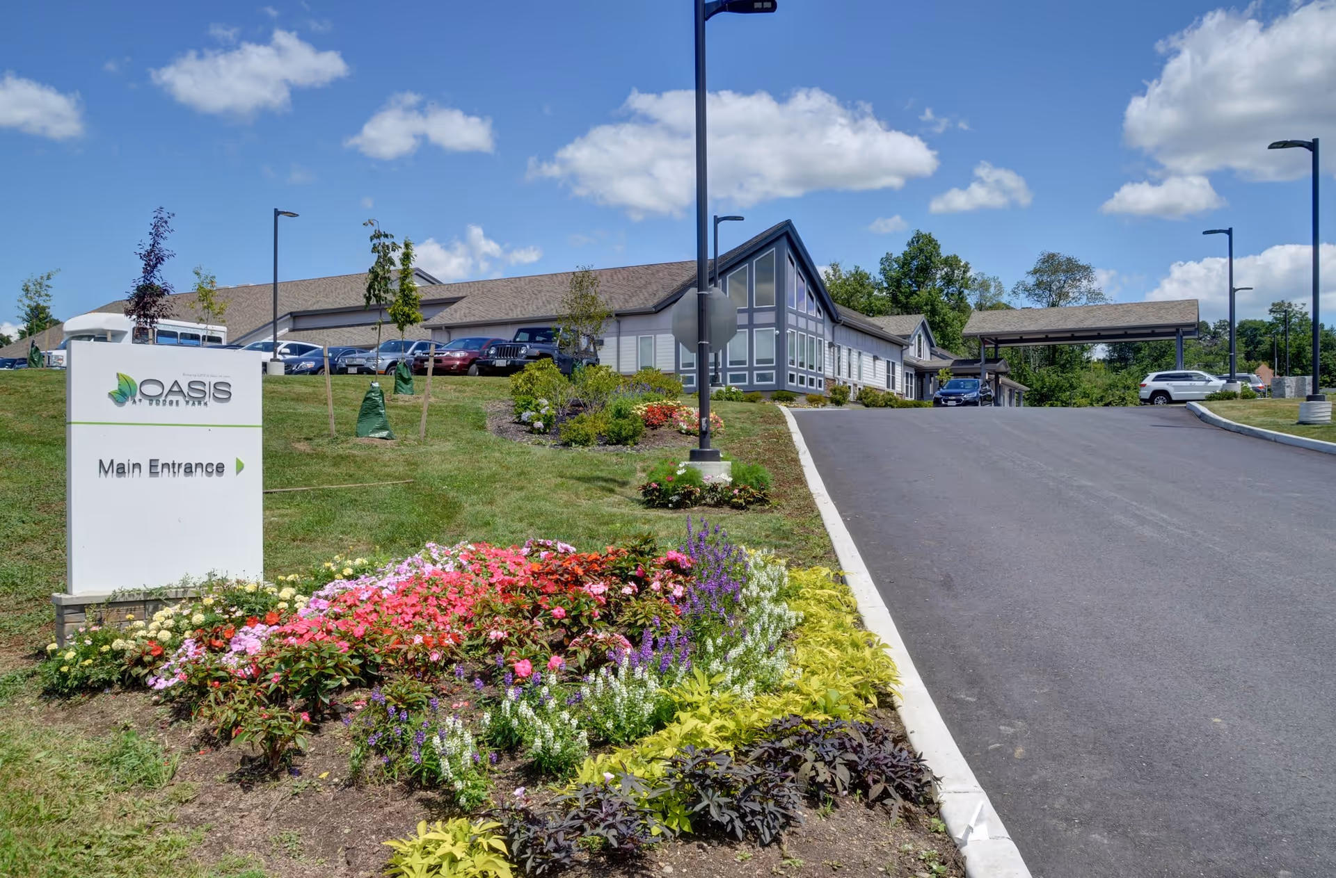 Exterior view of Oasis At Dodge Park facility with a paved driveway leading to the main entrance. The building has a modern design with large windows and a peaked roof. There is a landscaped garden with colorful flowers and greenery near a sign that reads 'Oasis At Dodge Park Main Entrance'. Several cars are parked near the building under a clear blue sky with scattered clouds.