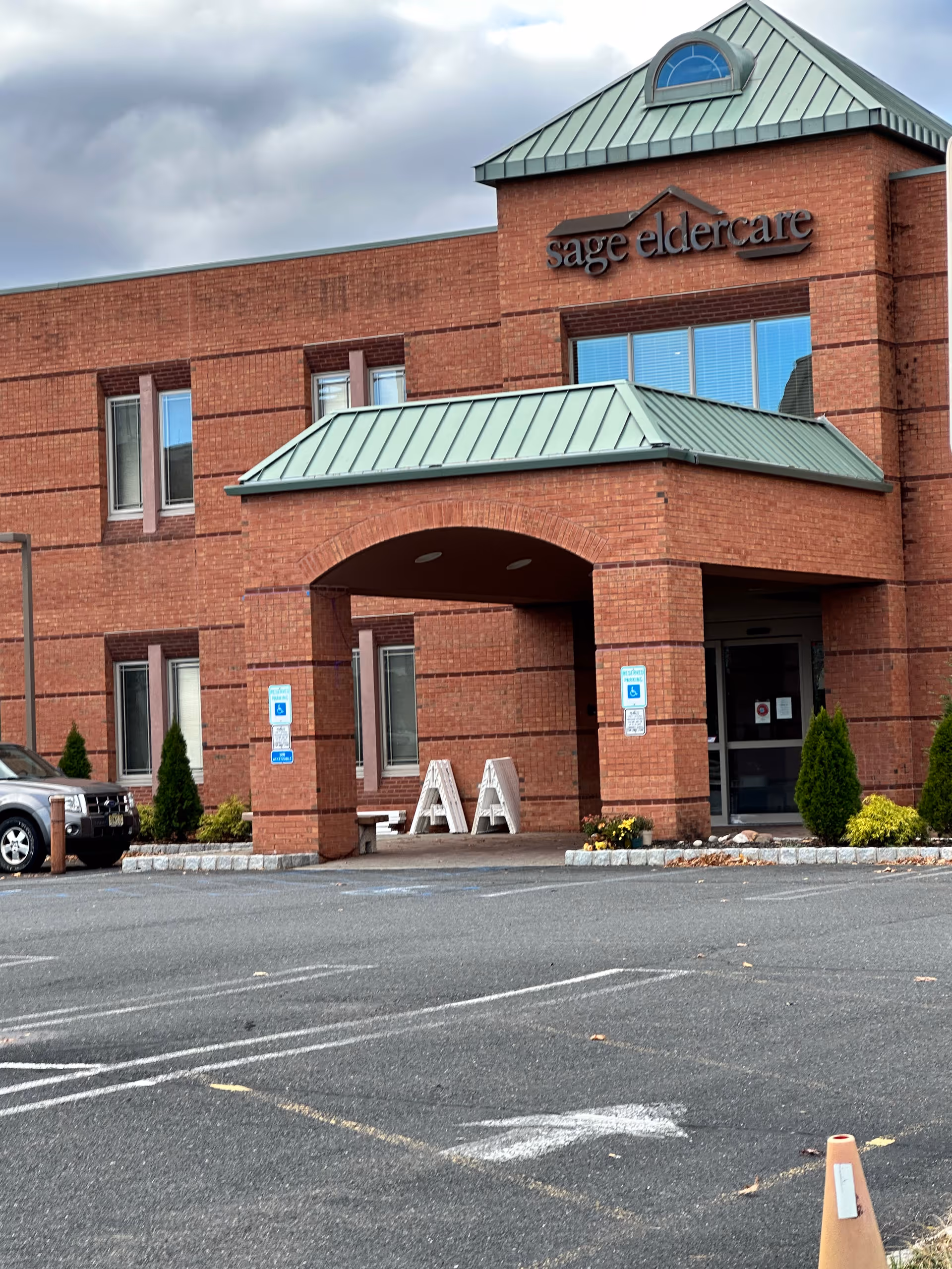Brick building labeled Sage Eldercare with a green-roofed entrance and parking lot.