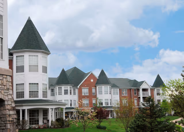 Red brick and white multi-story retirement community building with conical turrets and a landscaped lawn under a partly cloudy sky.