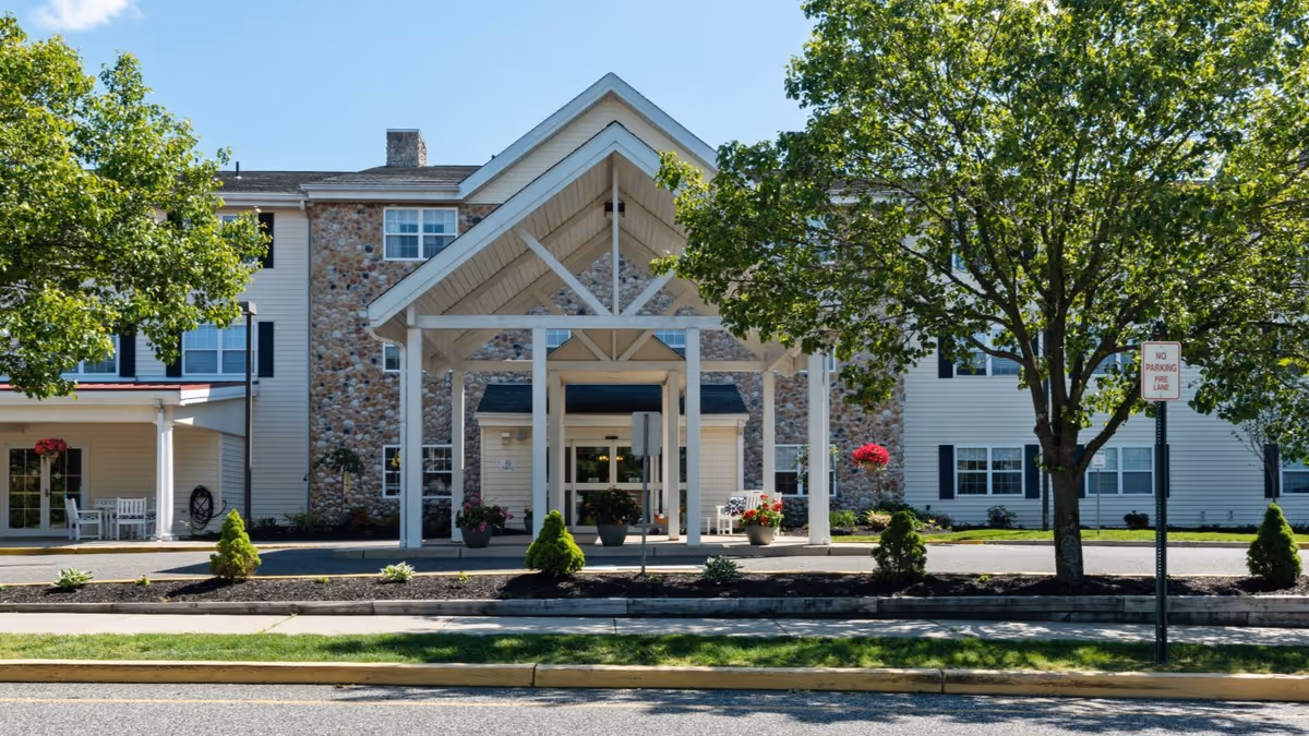 Front exterior view of a senior living facility building with a covered entrance, stone and siding facade, surrounded by trees and landscaping.