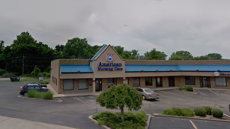 Front view of American Nursing Care facility with blue roof and parked cars.