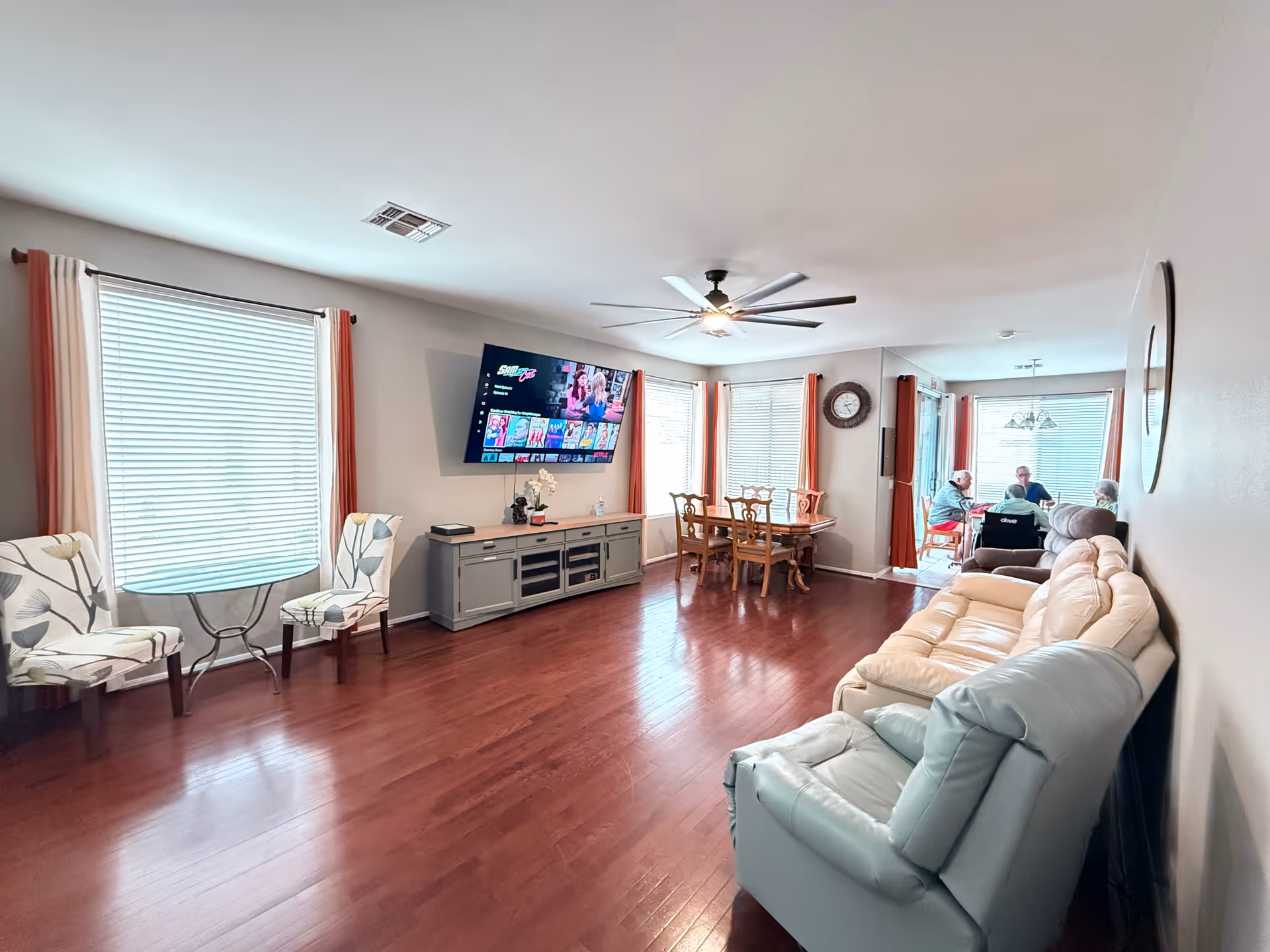 Spacious senior living room with a wall-mounted TV, sofas, chairs, and a dining area.