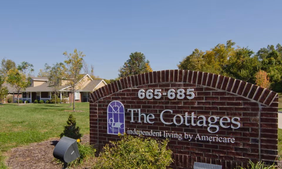 A brick sign for The Cottages independent living by Americare with the address 665-685, set in a landscaped area with grass, small bushes, and trees. In the background, there is a single-story residential building with a porch and a sloped roof under a clear blue sky.