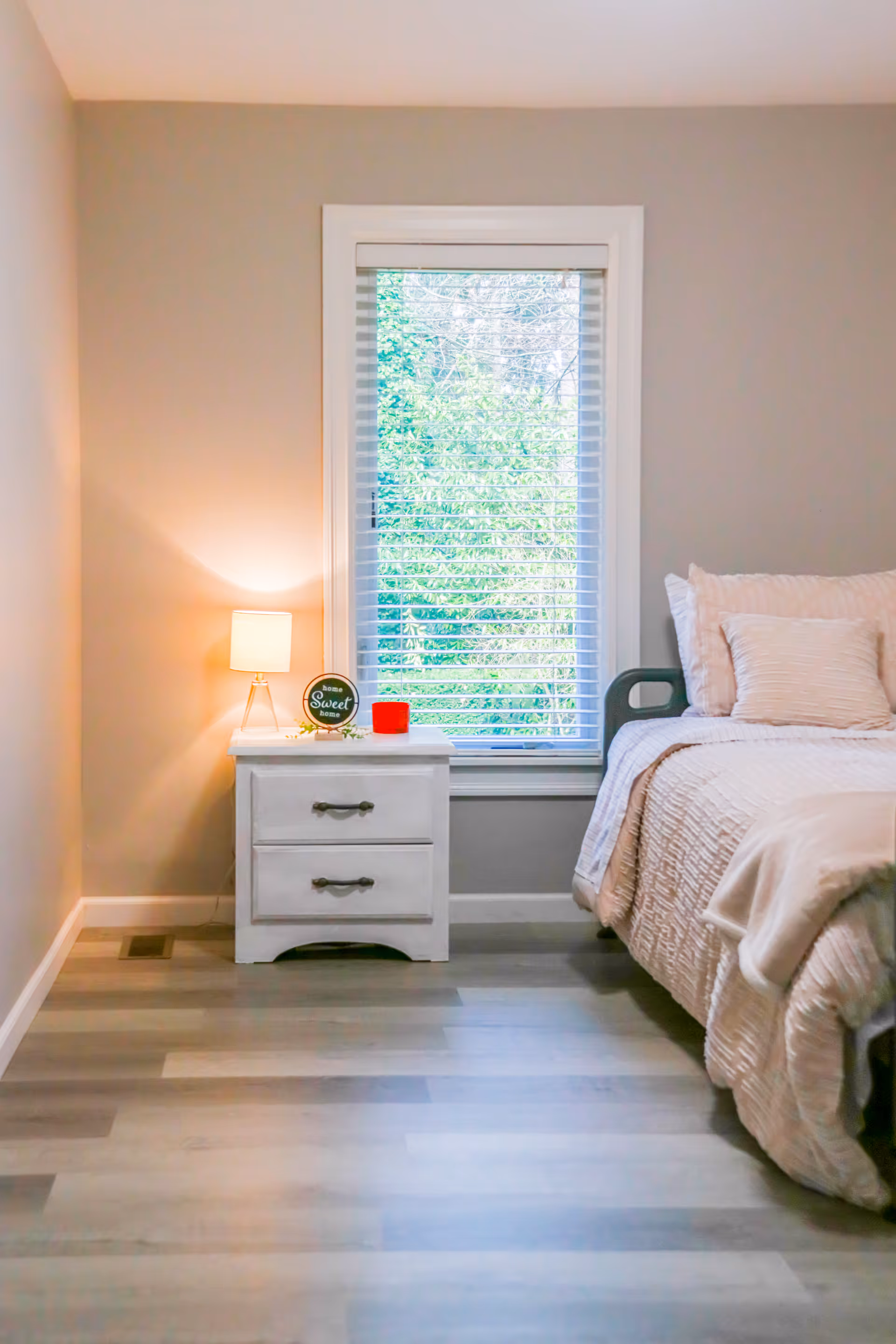 Light-filled bedroom with a nightstand, lamp, and bed by a window.
