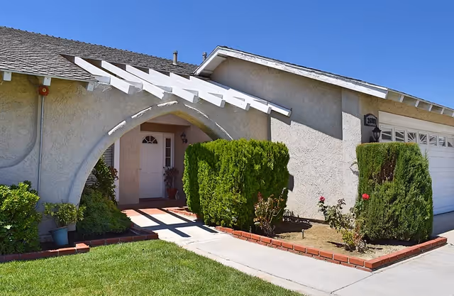 Residential care facility exterior featuring an arched entry, white stucco walls, and trimmed shrubs.