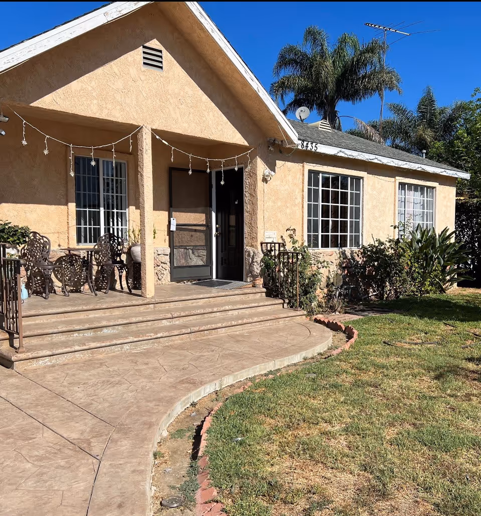 Front exterior view of a single-story beige stucco building with a small porch featuring metal chairs and a table. The building has large windows with white frames, a black door with a screen door, and a few steps leading up to the porch. There are palm trees and other greenery in the background under a clear blue sky.