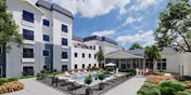 Outdoor courtyard area of a senior living facility with a swimming pool surrounded by lounge chairs and umbrellas. The multi-story building with white and gray exterior walls encloses the courtyard. Trees and landscaped bushes are visible around the pool area under a partly cloudy blue sky.