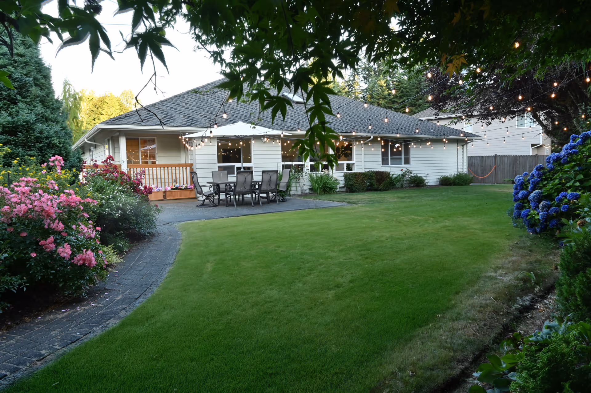 Single-story house with a cozy outdoor patio, string lights, green lawn, and flowering shrubs.