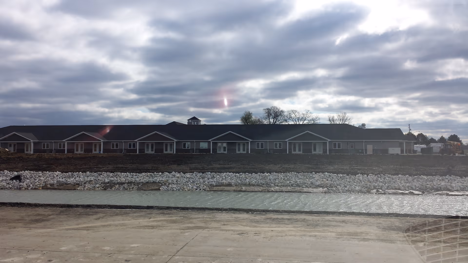 A long, single-story senior living facility sits along a rocky shoreline under cloudy skies.