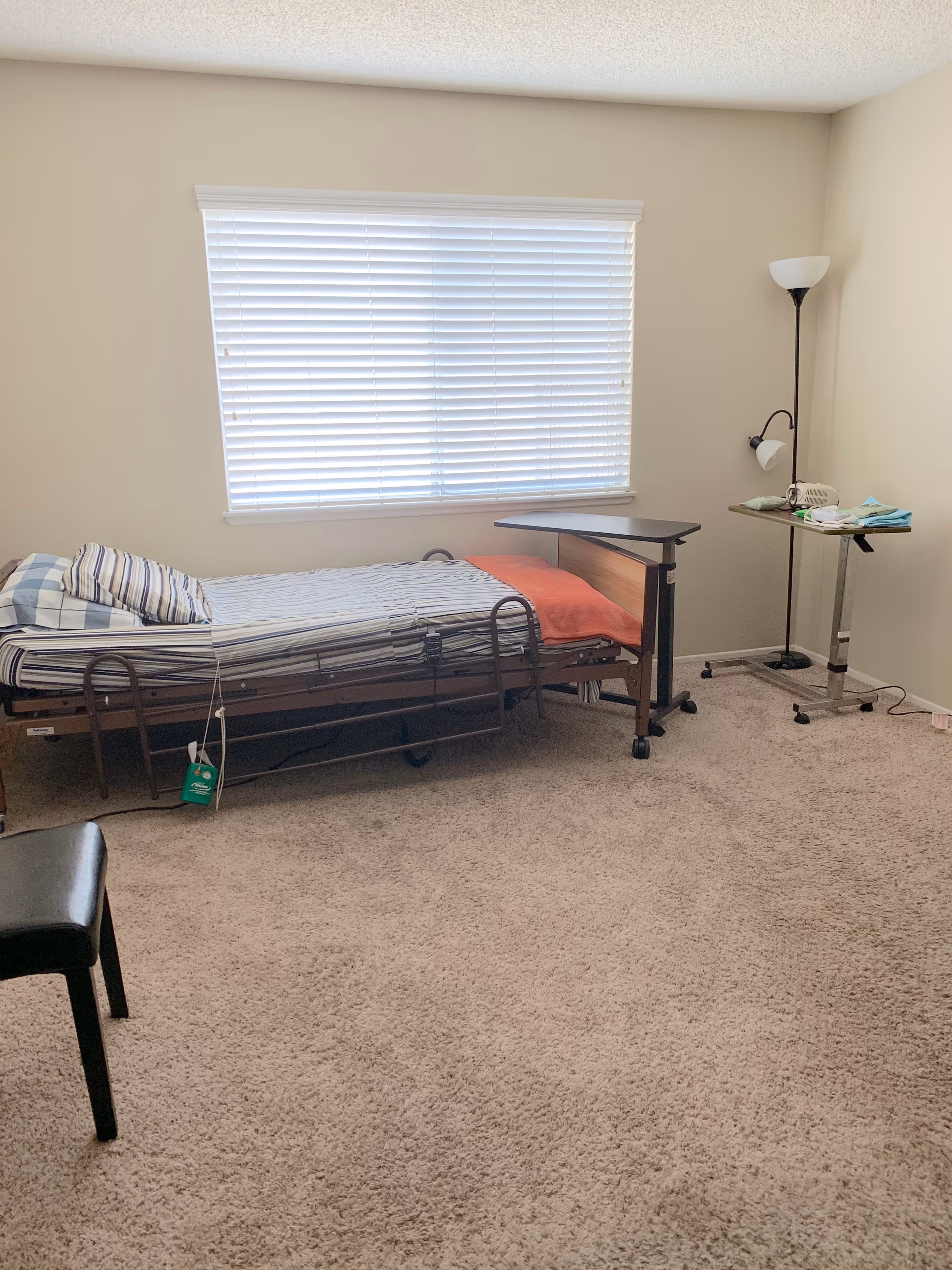 A furnished senior living bedroom with a single bed, desk, lamp, and window blinds.