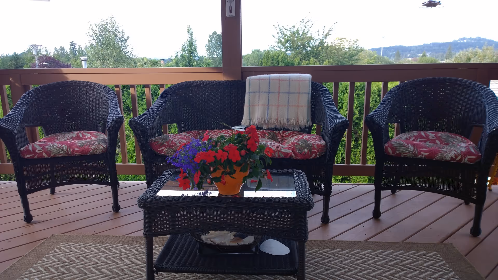 Wicker patio seating on a wooden deck with a coffee table, potted flowers and a blanket overlooking trees and distant hills.