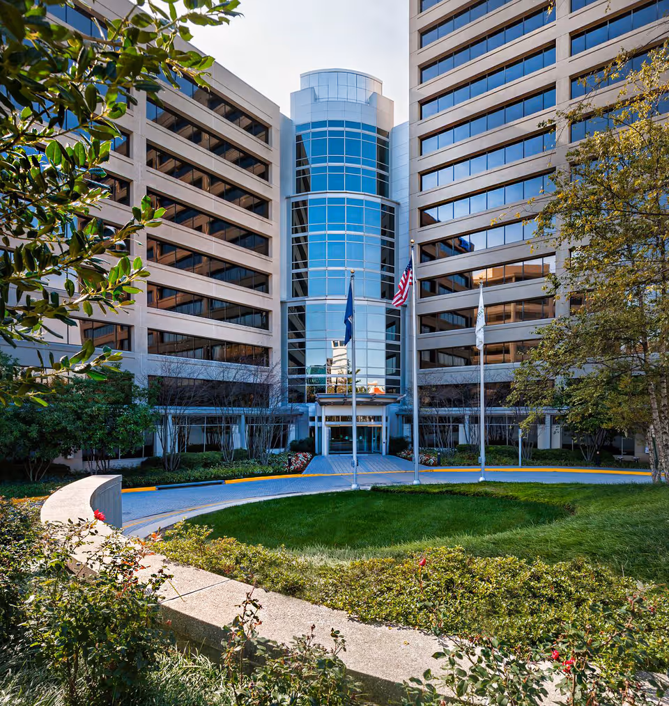 Modern multi-story building entrance with glass facade, flagpoles, and landscaped surroundings.