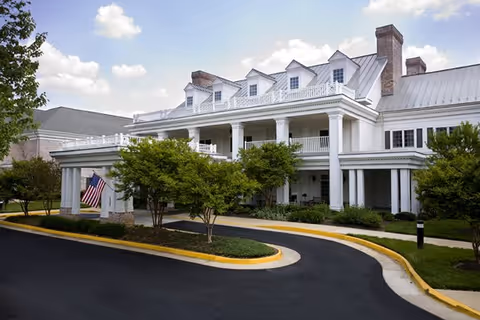 White multi-story senior living building with wraparound porch and well-kept landscaping.