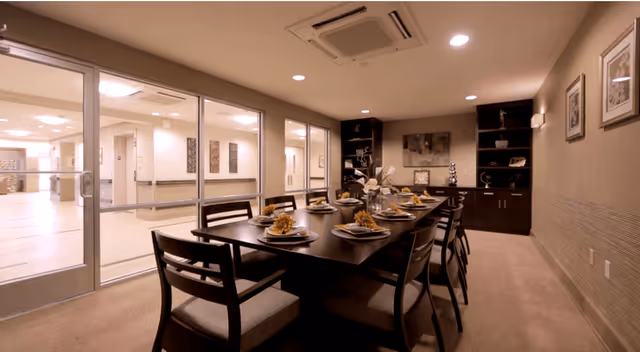 A dining room with a long dark wooden table set for a meal with plates, napkins, and decorative centerpieces. The room has beige walls, carpeted floor, and recessed ceiling lights. Large glass sliding doors open to a bright hallway or common area. There is a dark wooden cabinet with shelves and framed artwork on the walls.