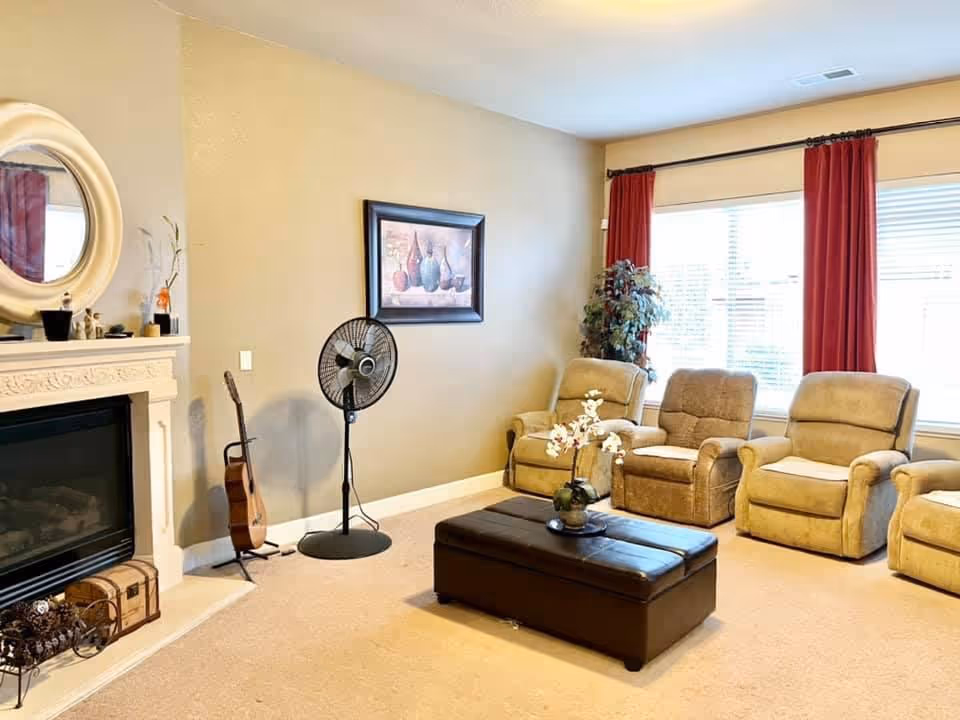 A cozy living room with four beige recliner chairs arranged in a row near a large window with red curtains. A dark brown ottoman with a flower vase on top is placed in the center of the room. To the left, there is a white fireplace with decorative items on the mantel, a round mirror above it, a standing fan, and a guitar resting against the wall. A framed painting hangs on the beige wall above the fan.