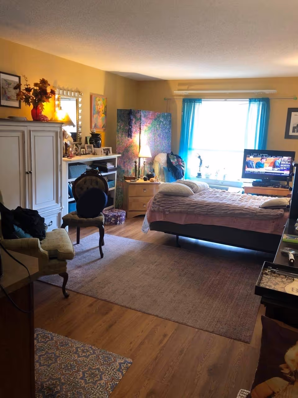 A cozy bedroom with a bed covered in a pink quilt and pillows, a TV on a stand showing a news program, a window with blue curtains letting in natural light, a wooden floor with a large area rug, an armchair, a small table with a lamp, and various decorative items including framed pictures and a colorful room divider.