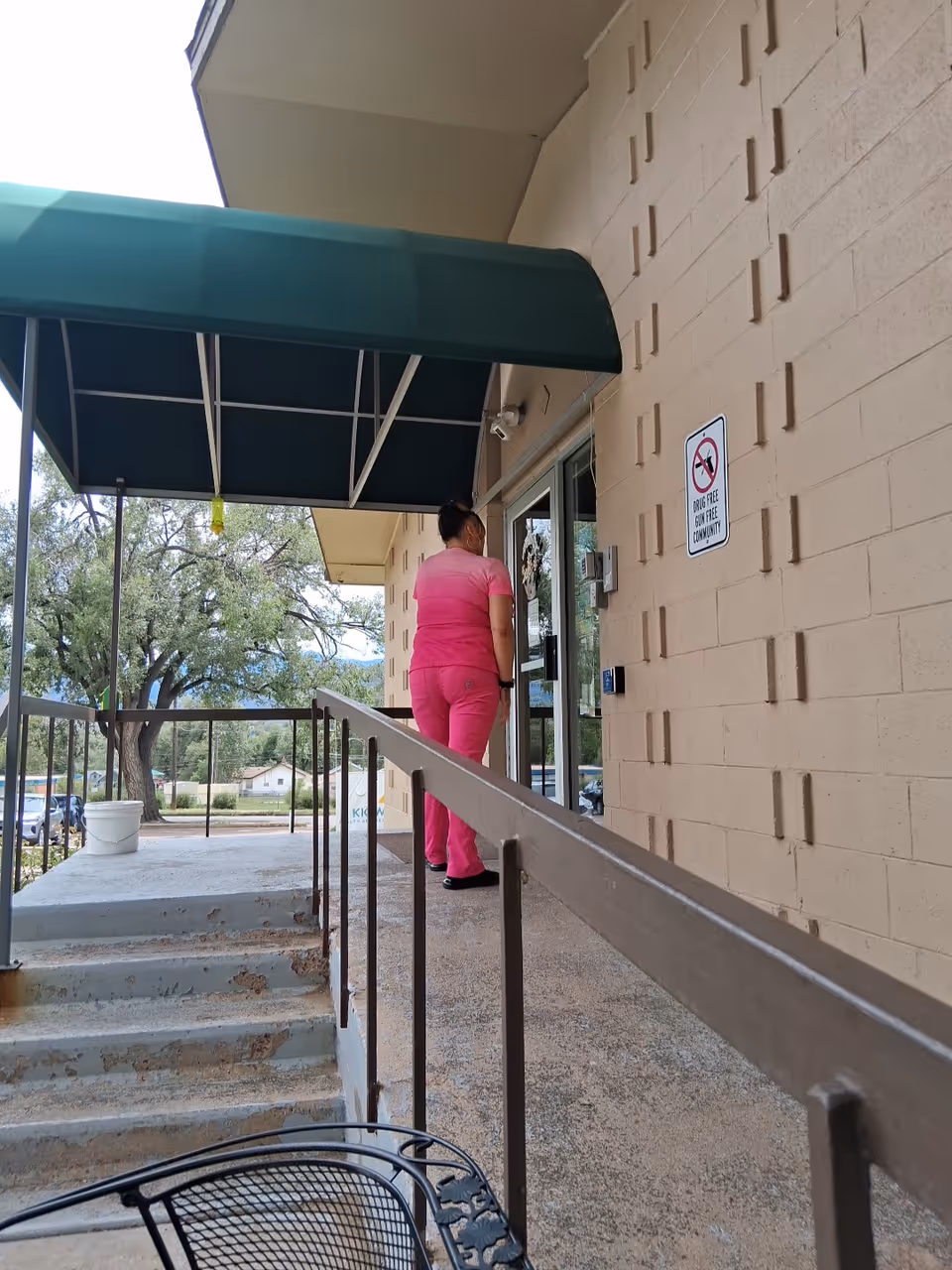 A person in pink scrubs enters a building through a glass door under a green awning.