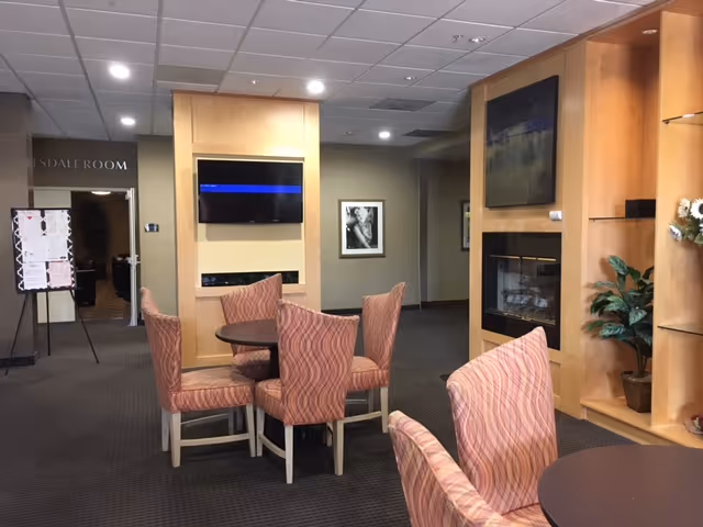 Senior living lounge with pink upholstered chairs around tables, a fireplace, and wall TVs.