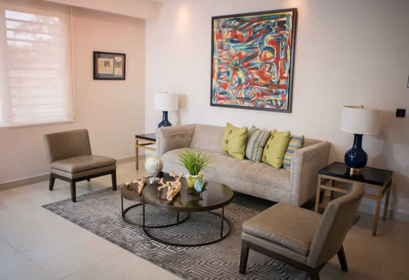 A modern living room with a beige sofa adorned with green and striped cushions, two brown leather chairs, a round dark wood coffee table with decorative items including a small globe, plant, and driftwood pieces, two side tables each with a blue lamp, a large colorful abstract painting on the wall, and a window with a white blind.