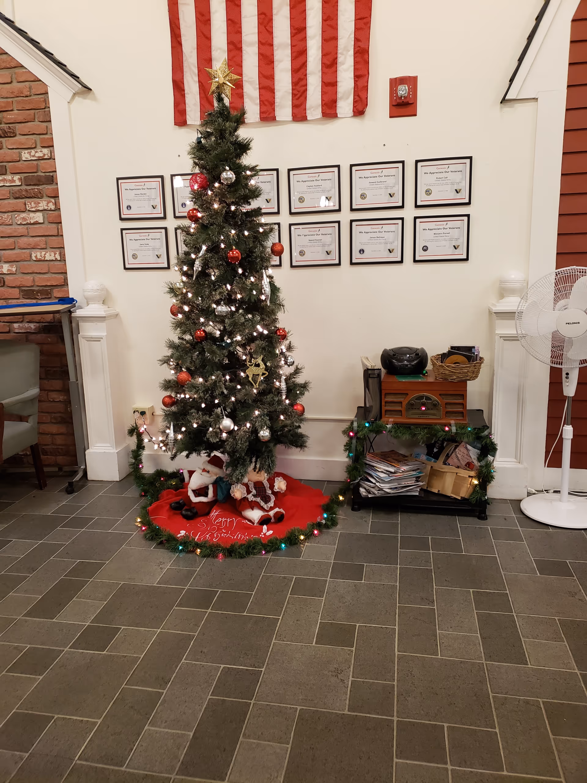 Christmas tree with lights and ornaments in a hallway with certificates on the wall.
