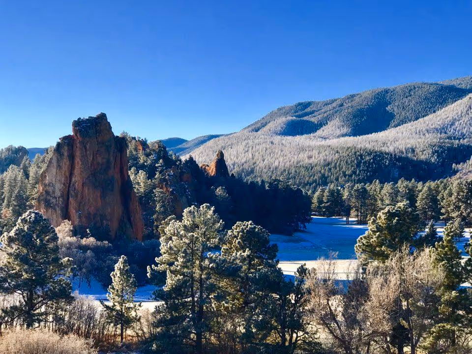 Snowy mountain landscape with rock formations, pines, and clear blue sky.