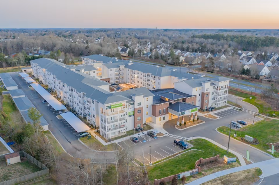 Aerial view of Sage Highland Creek, a large multi-story senior living facility with a covered entrance and parking areas. The building is surrounded by trees and residential houses in the background.