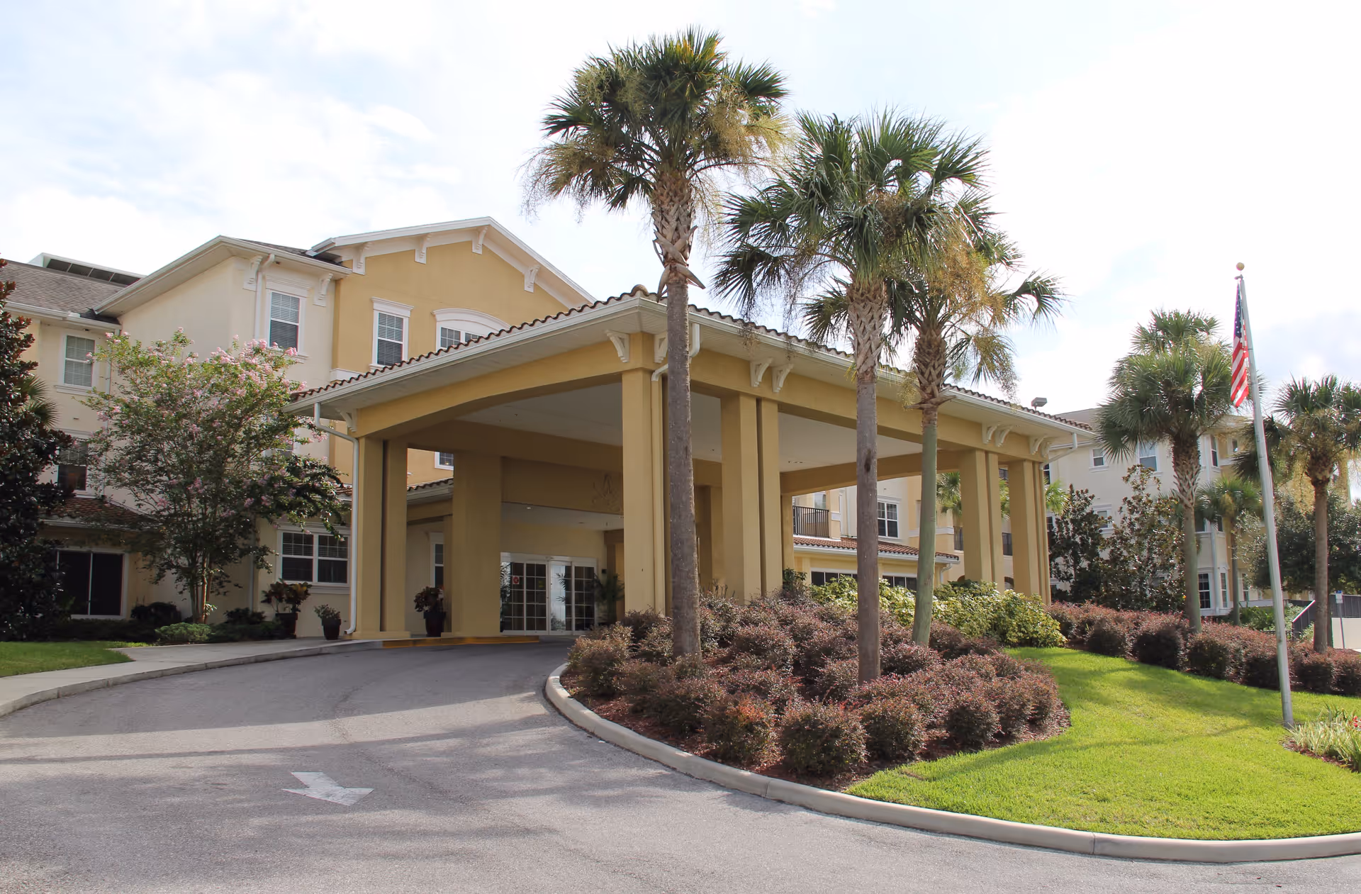 Front entrance of a multi-story senior living building with a covered porte-cochère, palm trees, and landscaped grounds.