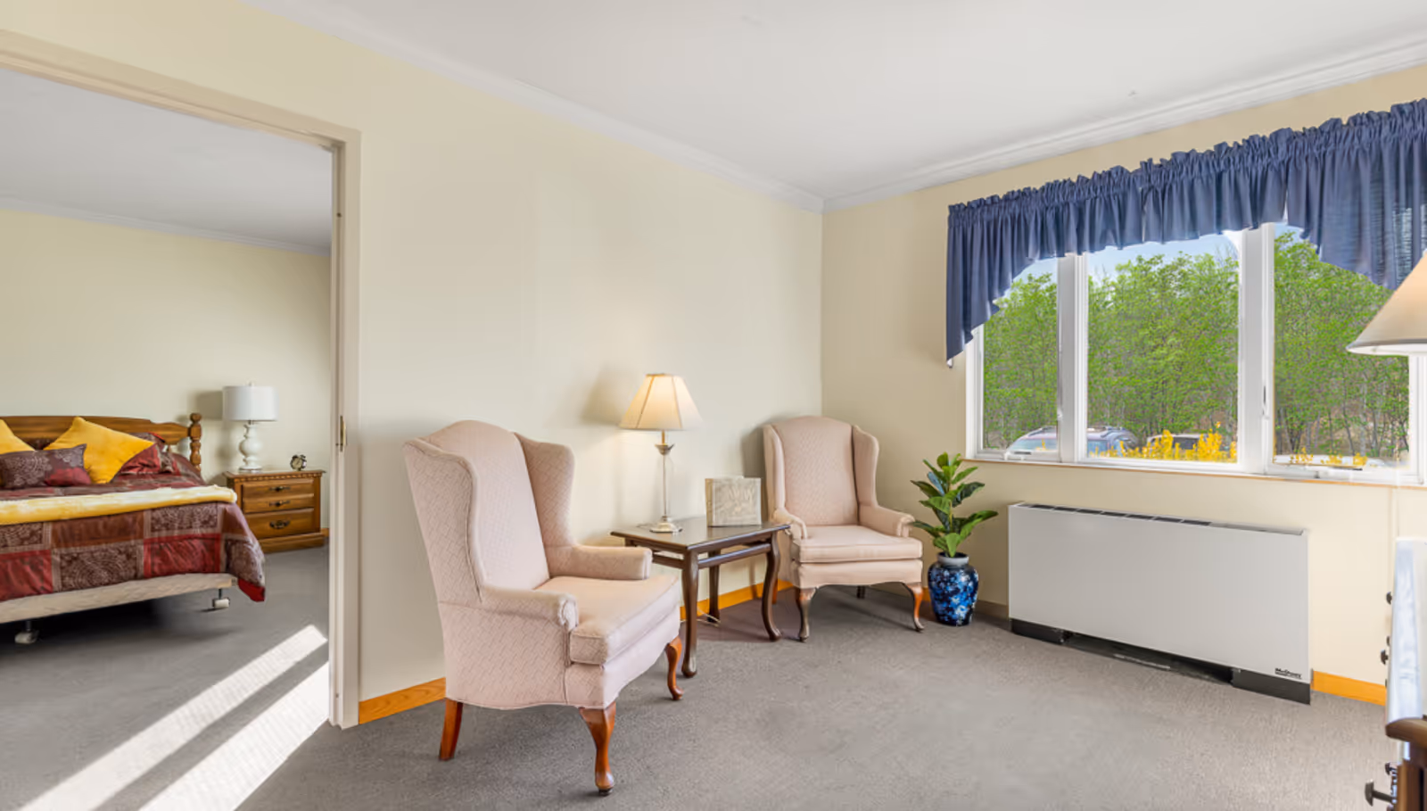 A cozy senior living room with pink upholstered chairs, a small table, lamp, and large window.