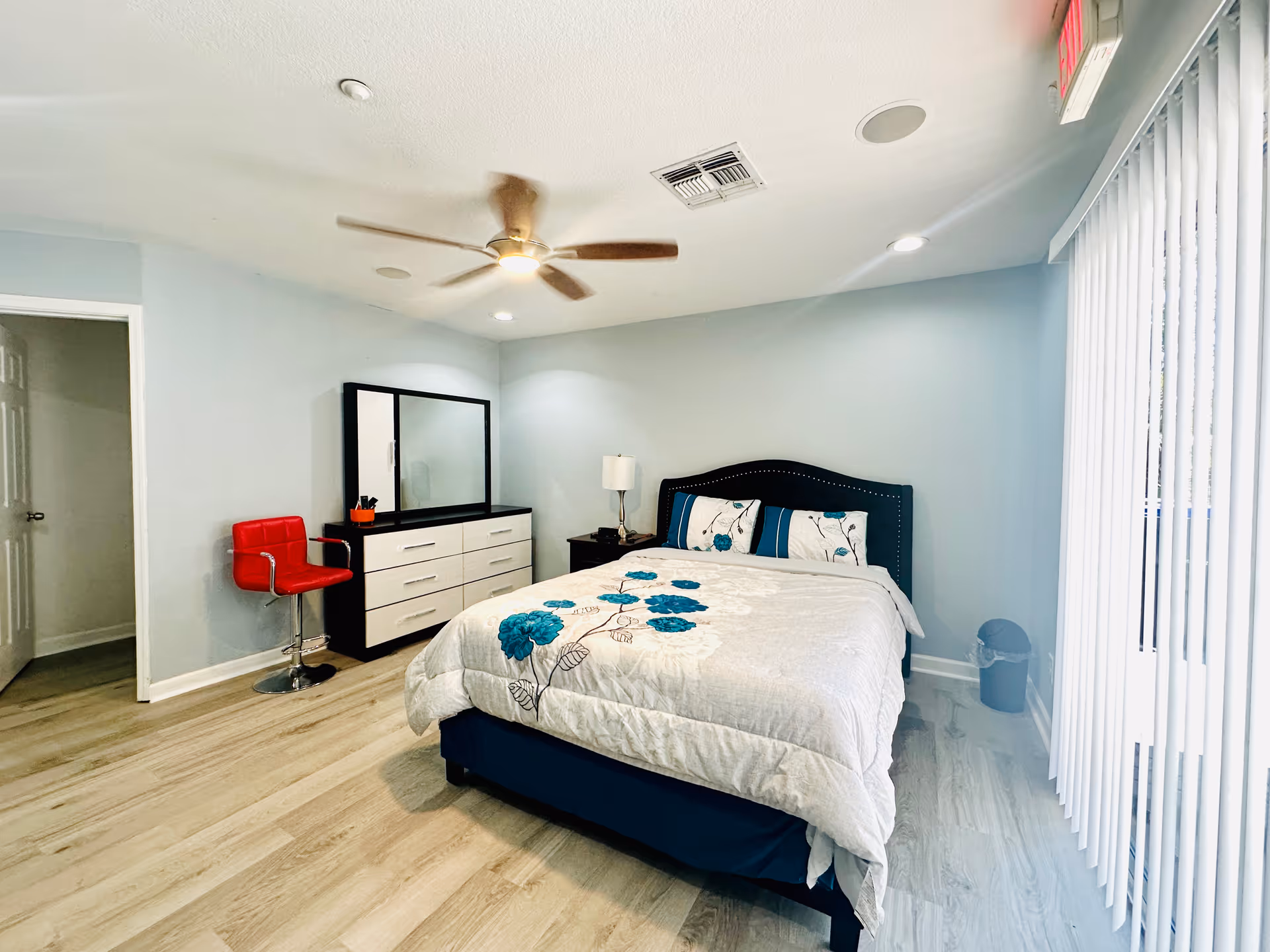 Bright modern bedroom with a bed, dresser, mirror, ceiling fan, red chair, and vertical blinds.